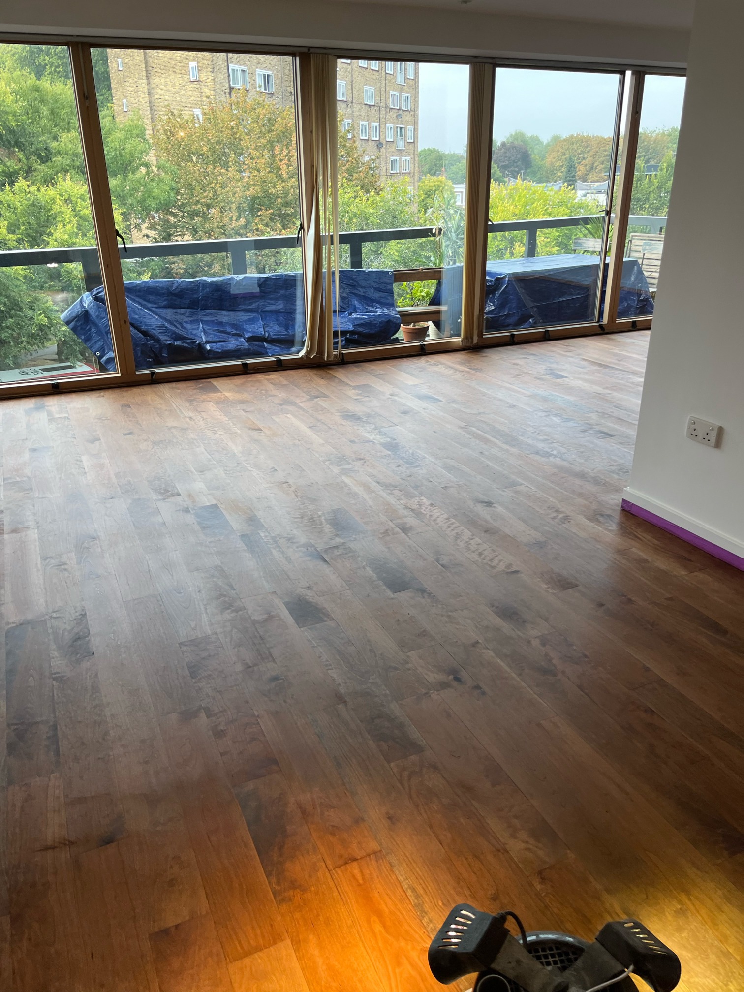 Living room with a freshly coloured hardwood floor in front of floor-to-ceiling windows, with a floor machine at the bottom of the frame