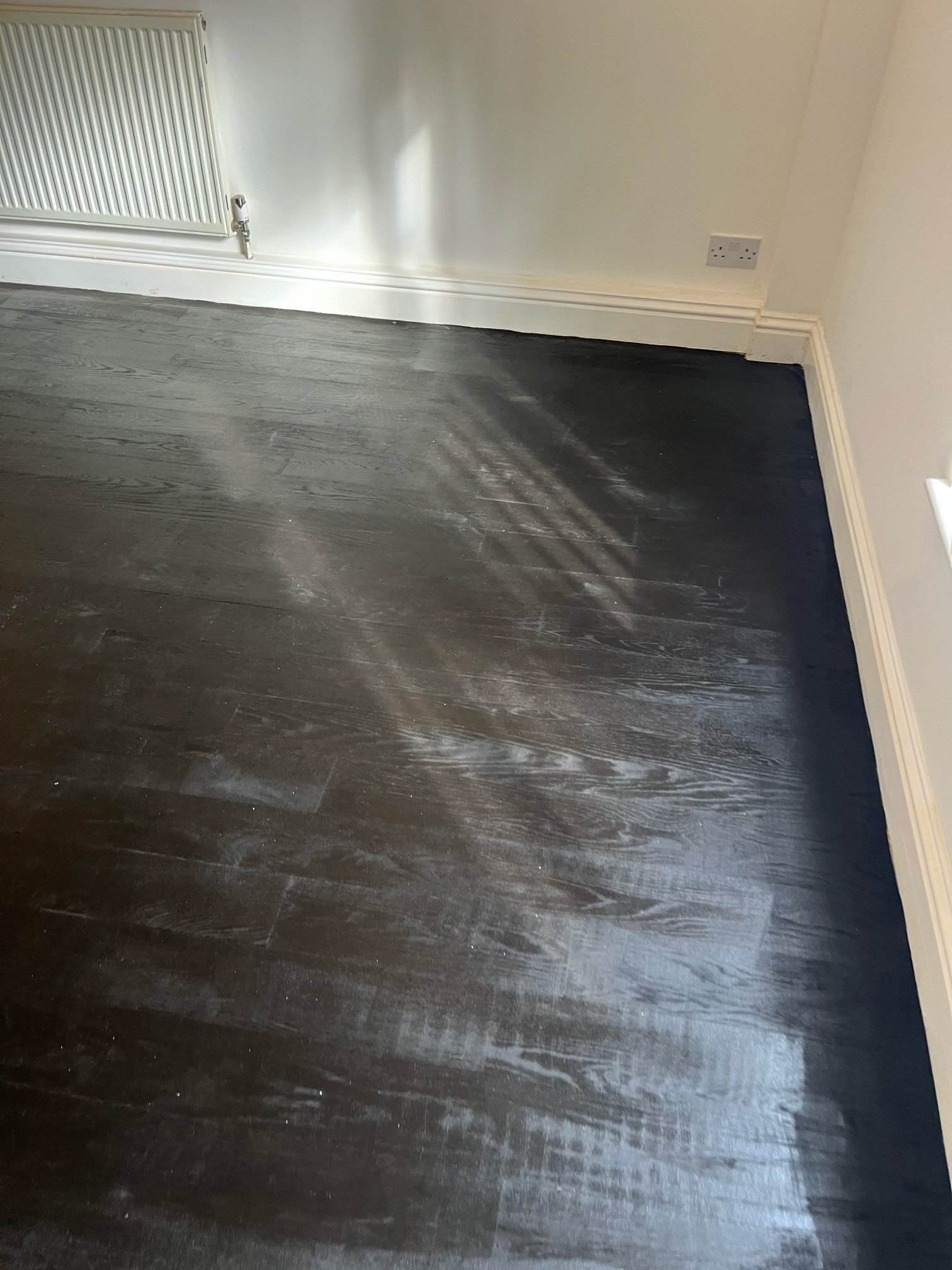Corner of a room with dark stained oak flooring, a white radiator and a double socket, with sunlight showing pale, patchy areas across the floor.