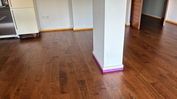 Richly coloured hardwood floor around a white column with purple tape at its base, with kitchen units behind