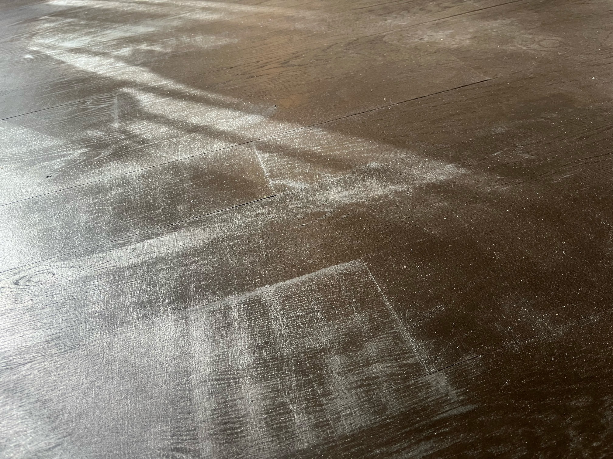 Low-angle view of dark stained oak flooring with a whitish haze over several boards, lit by light from a window.