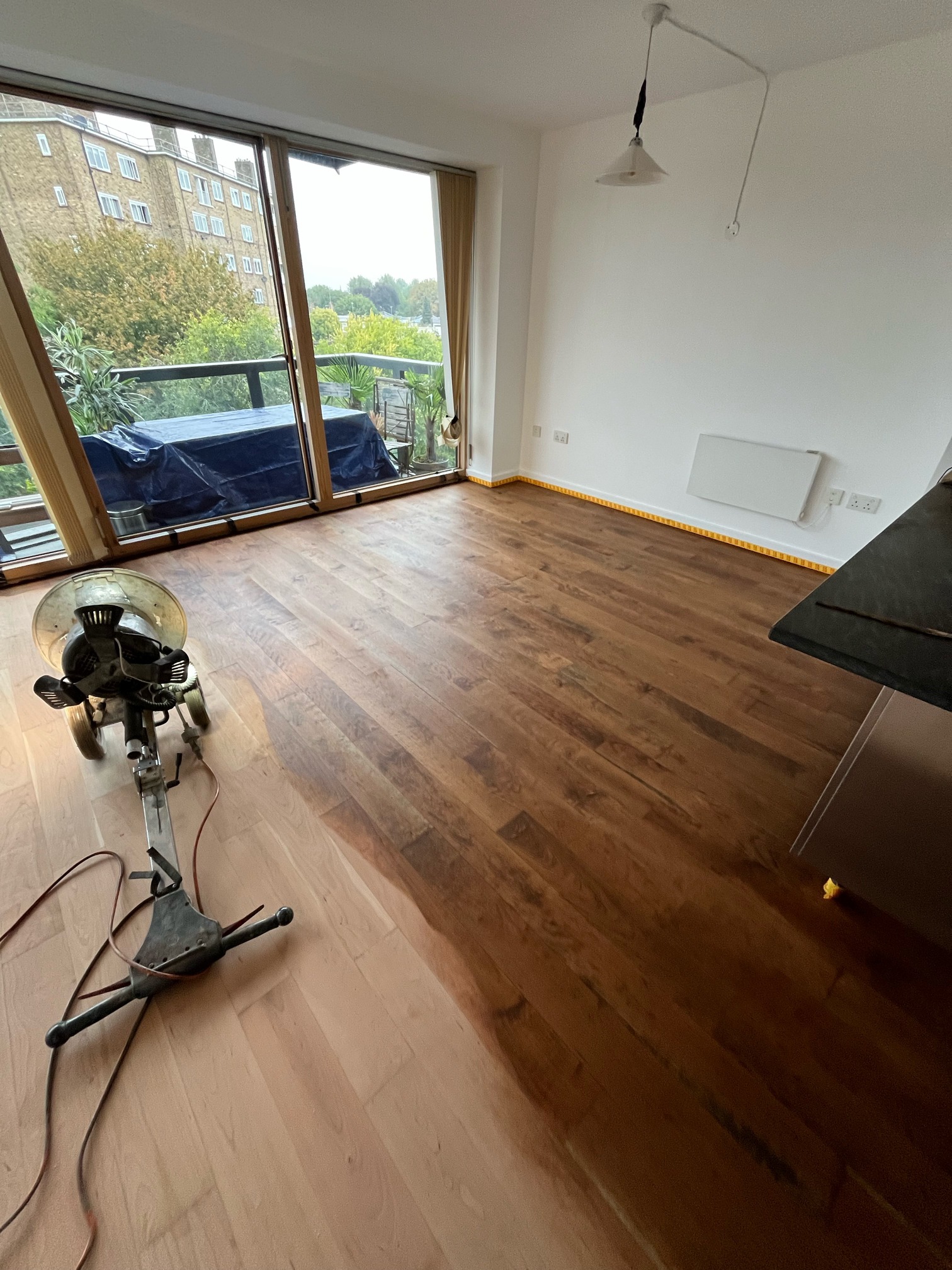 Hardwood floor in front of a balcony window, dark stained on one side and bare sanded wood on the other, with a floor machine and cable