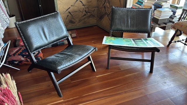 Two black leather lounge chairs on a worn hardwood floor beside stacked books and a folding screen