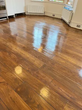 A freshly varnished wooden floor with a glossy reflective finish in a bright room, showing rich brown wood grain and natural light from nearby windows.