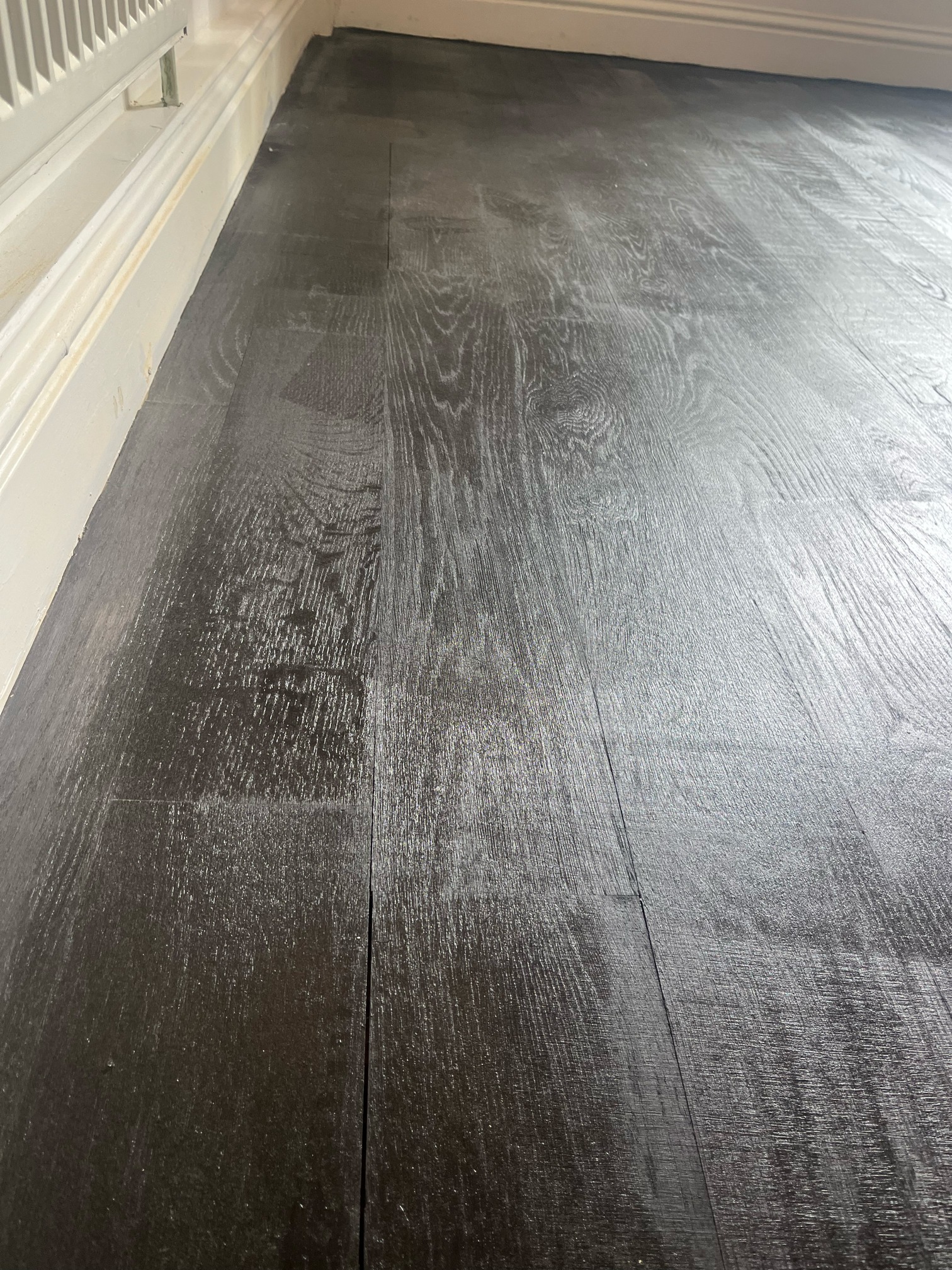 Dark stained oak boards running along a white skirting board under a radiator, with hazy, uneven colour and an open joint between two boards.