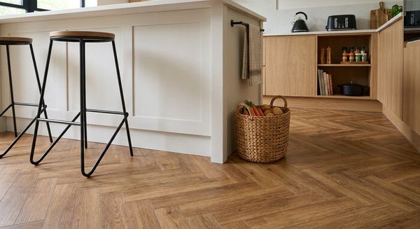 Kitchen with oak-effect herringbone LVT flooring, a white panelled island, black metal bar stools and a wicker basket of vegetables
