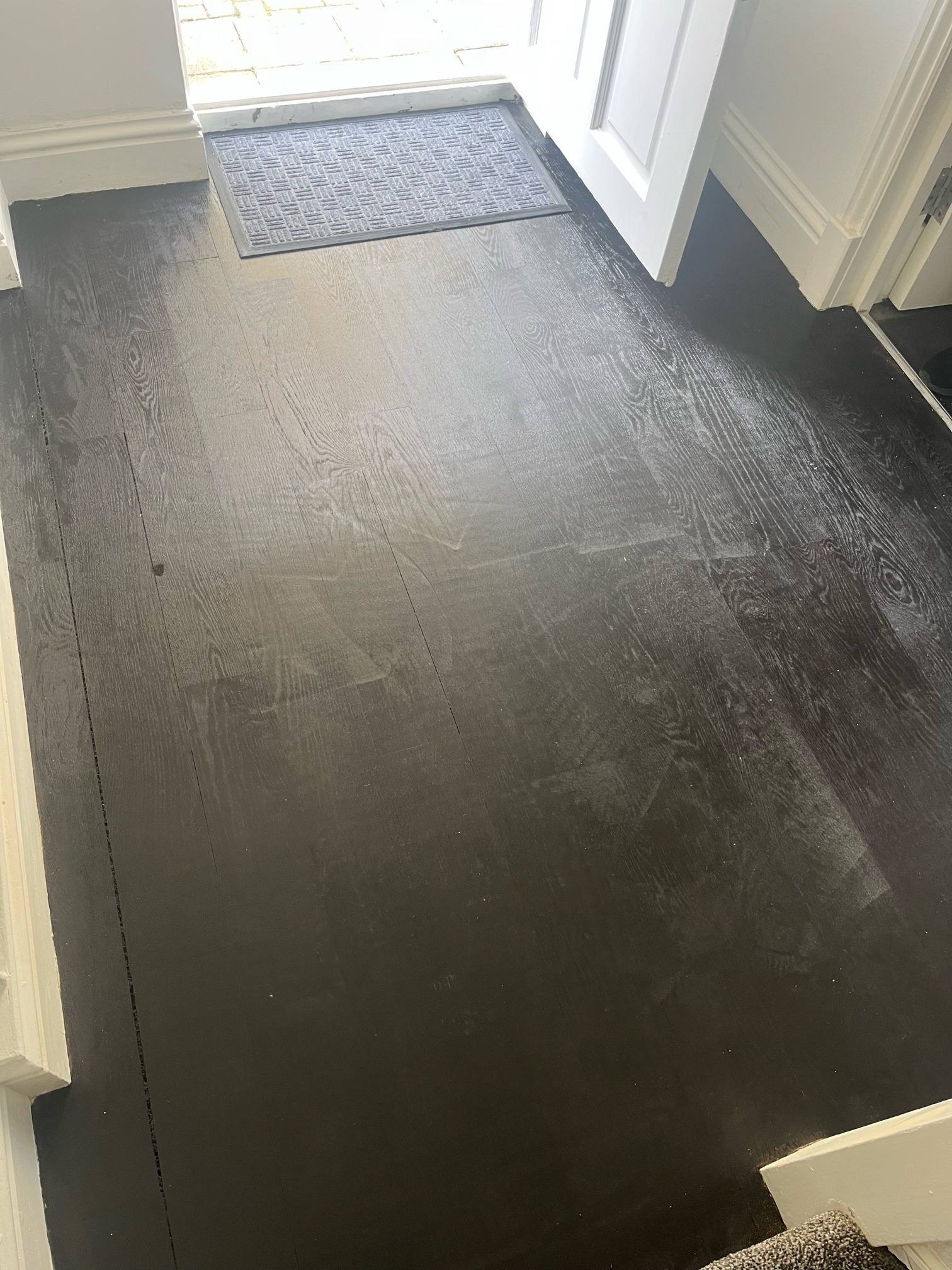 Hallway with dark stained oak flooring leading to an open front door and doormat, with a visible gap between two boards.