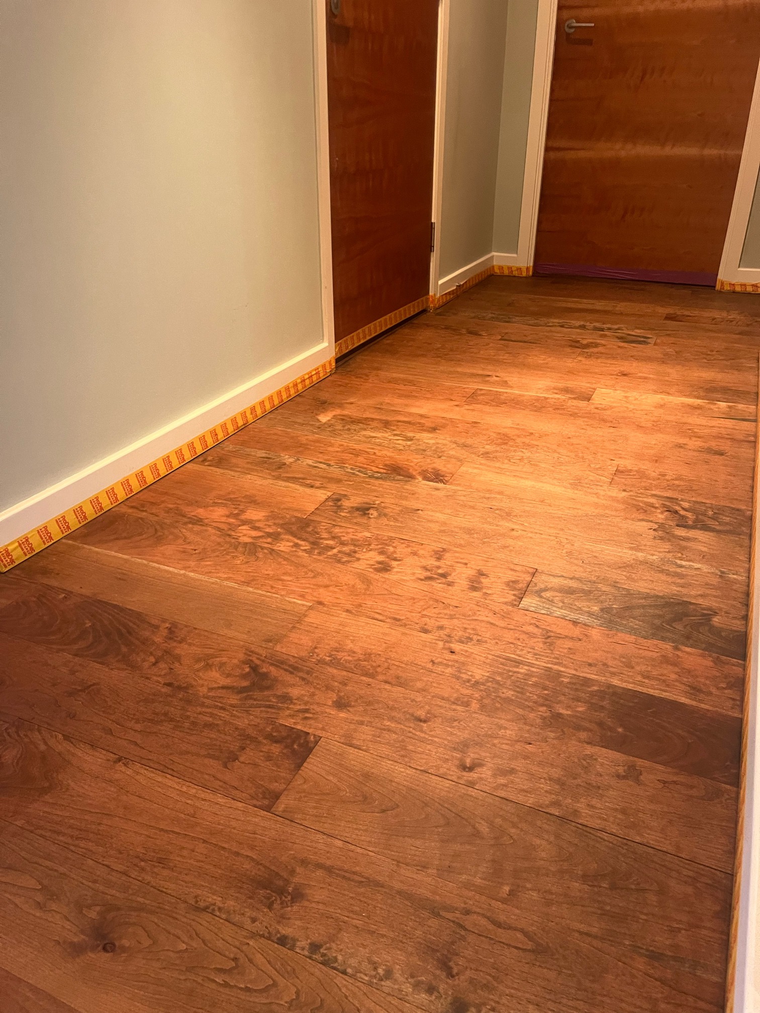 Hallway hardwood floor with warm orange-brown tones and visible grain, with masking tape along the skirting