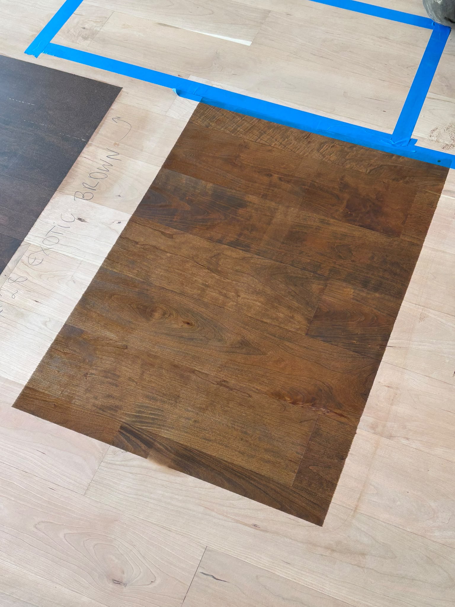 Close view of a mid-brown stain sample applied to bare sanded hardwood floorboards, with a darker sample at the left edge