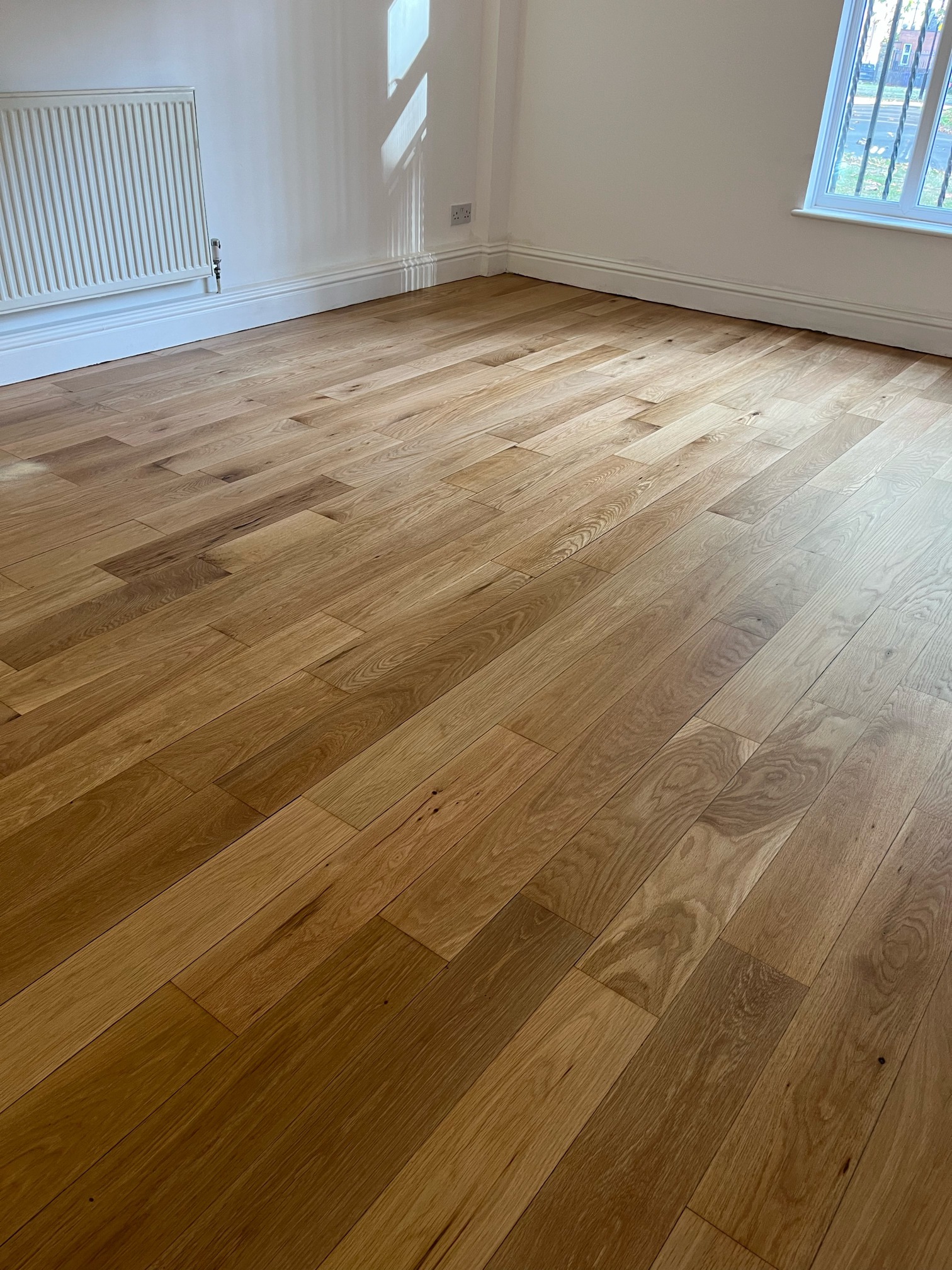 Empty room with natural oak plank flooring, a white radiator and a window with railings outside.