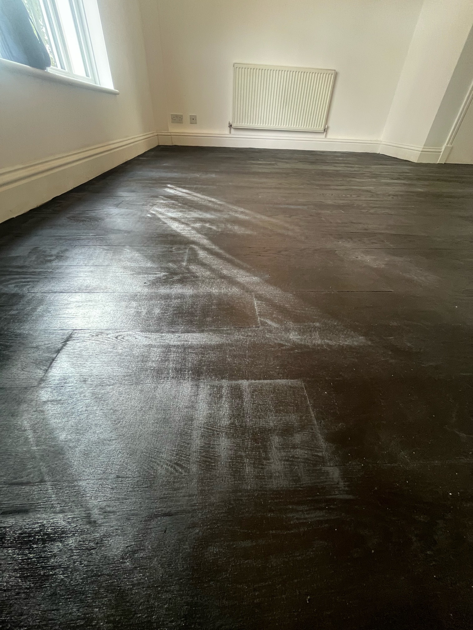 Dark stained oak floor with streaky, hazy patches catching the light, in a room with a radiator and window.