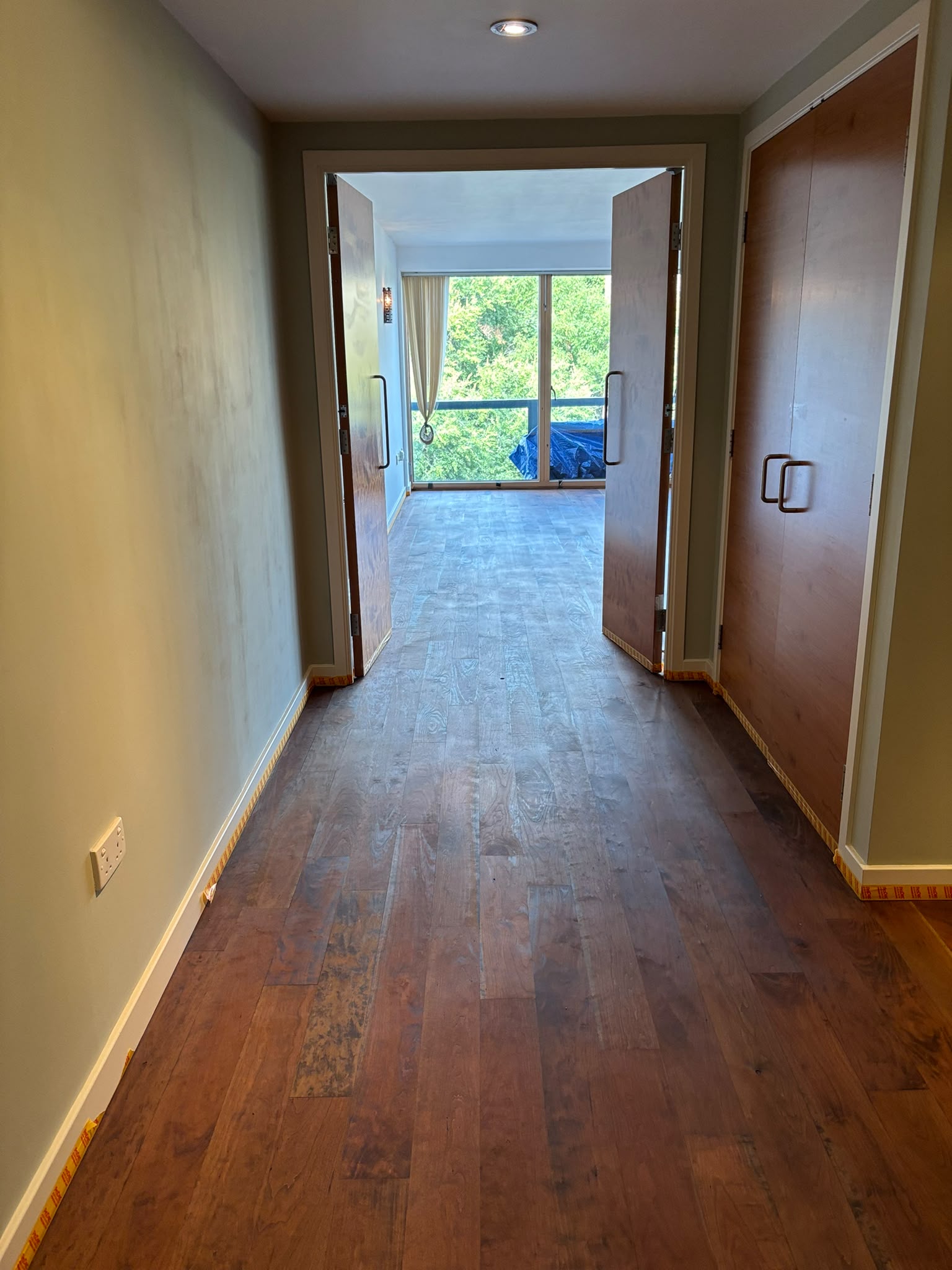 Hallway leading to double doors and a bright living room, with hardwood floor and yellow tape along the skirting boards