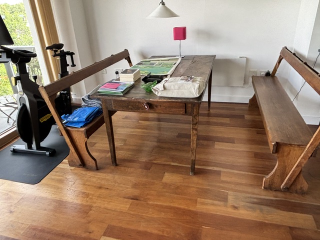 Worn hardwood floor around a wooden dining table and two antique benches, with an exercise bike by the window