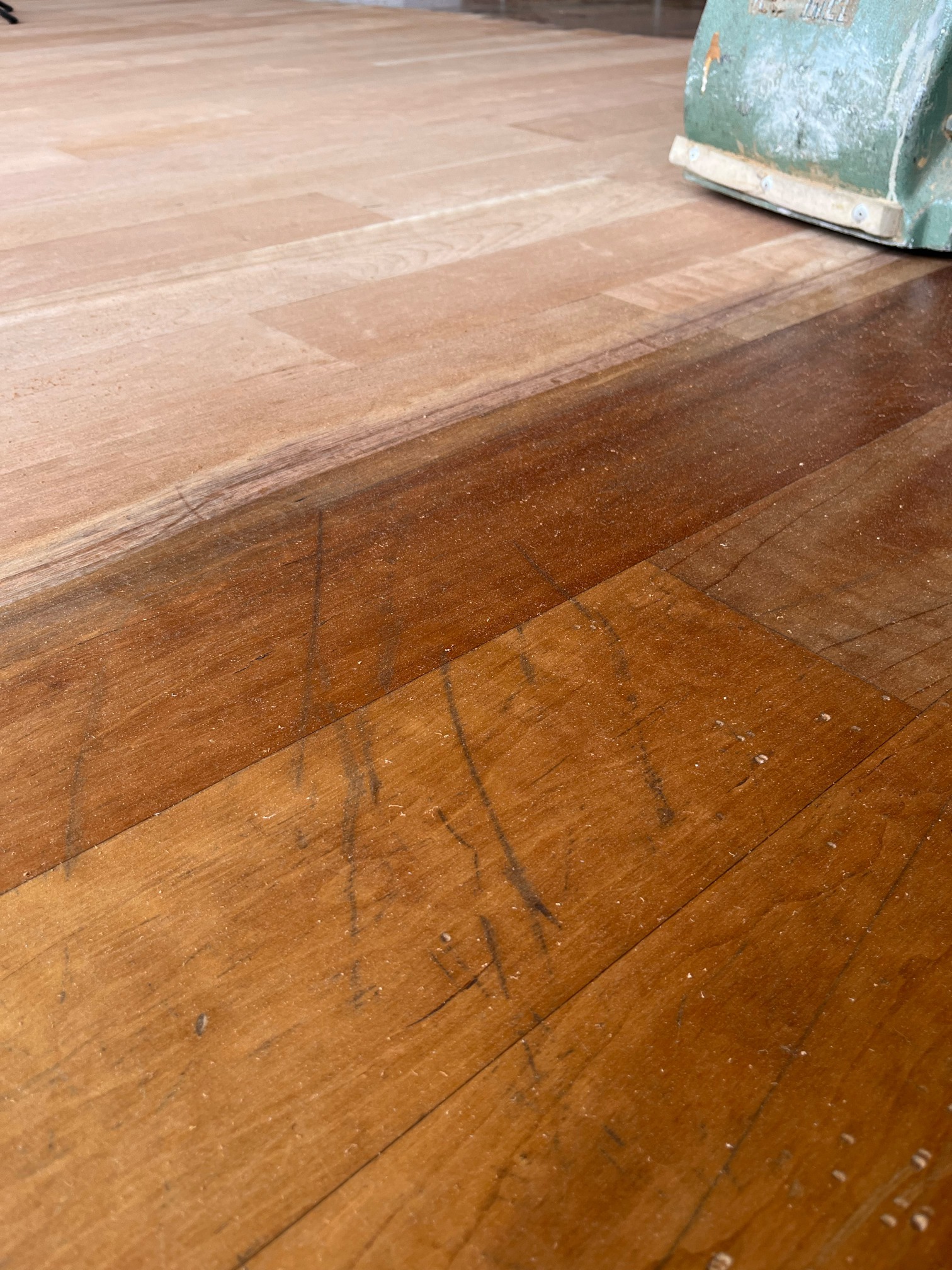 Hardwood floor with scratches in the old finish beside a freshly sanded area and a sander in the corner