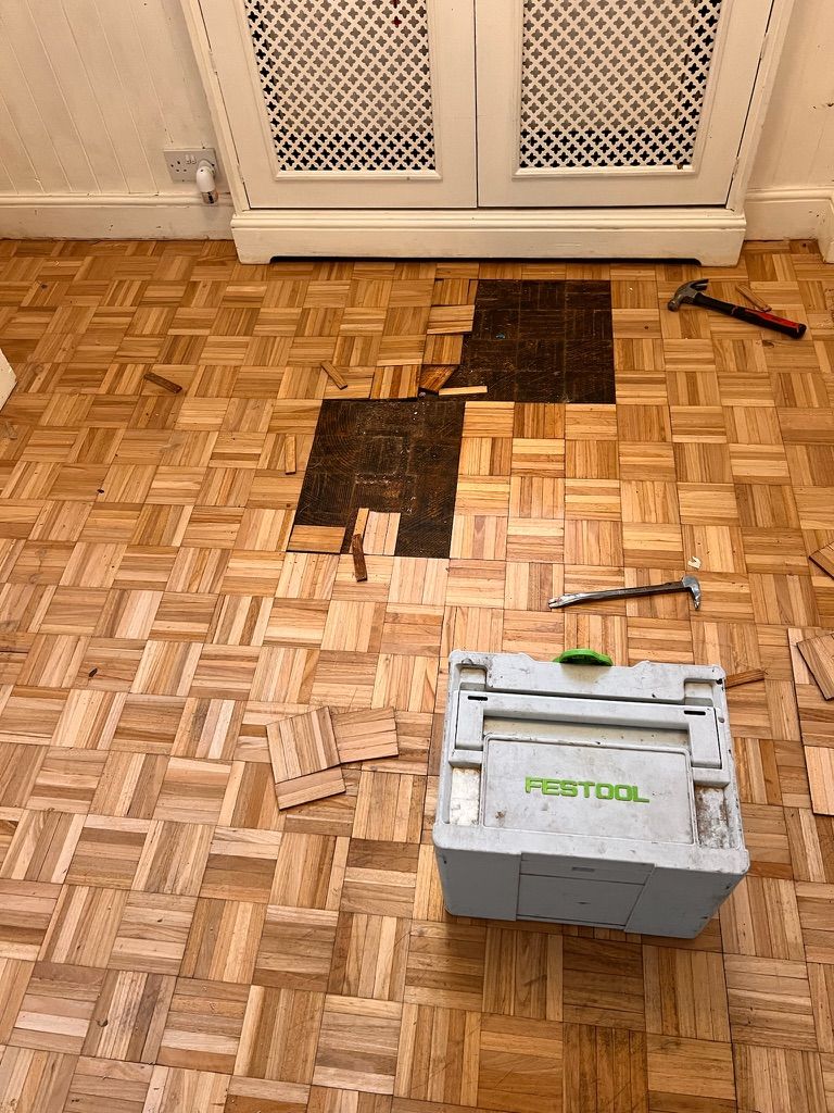 Parquet floor mid-repair with damaged wood blocks removed, replacement pieces and tools laid out on the floor.