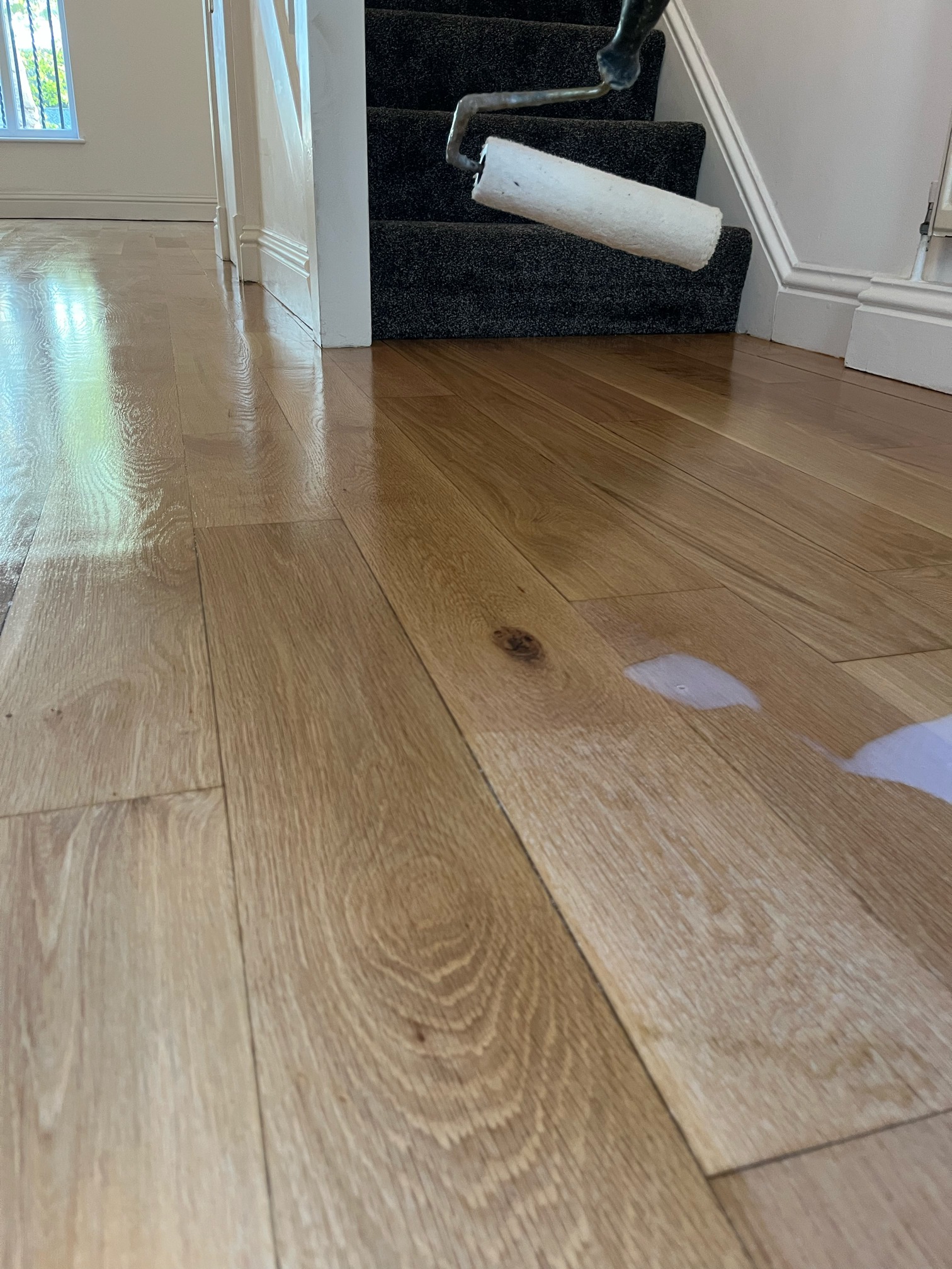Roller applying a clear finish to natural oak flooring at the foot of a carpeted staircase, half the floor already glossy.
